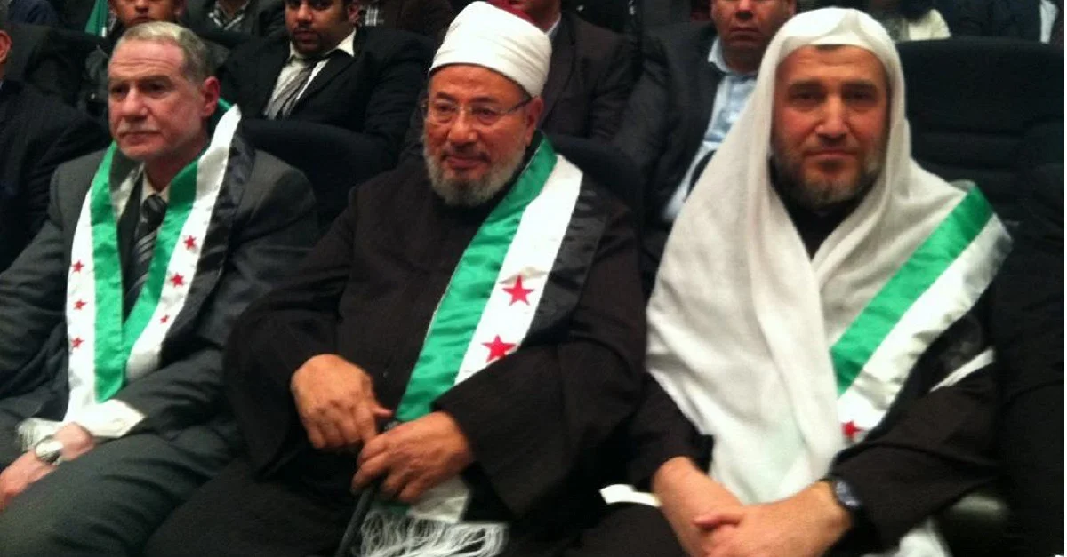 Bild (Yusuf al-Qaradawi [mitte] mit Flagge der syrischen Rebellen, 2013): Bilal Dweik (بلال الدويك) via Wikimedia Commons [CC BY-SA 3.0]
