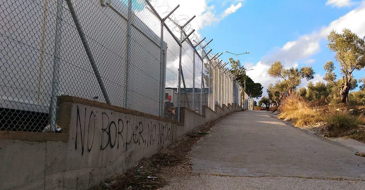 Bild (Mauer des Aufnahmelagers Moria): Cathsign [CC BY-SA 4.0], via Wikimedia Commons (Bild zugeschnitten)
